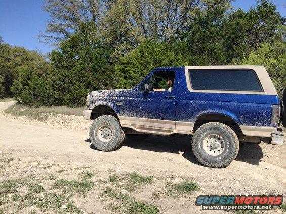 Blue 1996 Ford Bronco covered in mud, seen from the side on a dirt road
