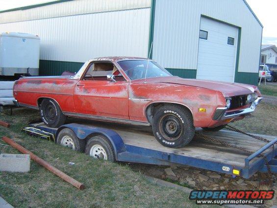 Red 1970s pickup truck on a blue trailer, parked on grass in front of a white building