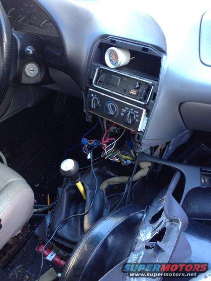 Car interior showing dashboard with radio removed, exposed wires, and an aftermarket gauge installed above the radio slot