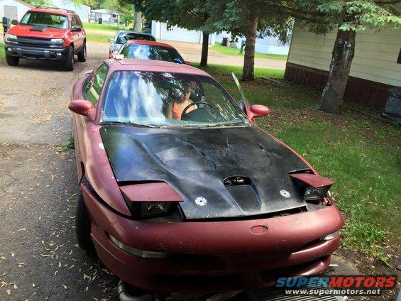 Maroon 1995 Ford Probe with black hood and raised headlights parked on gravel.
