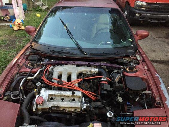 Maroon car engine bay with visible engine components and wiring. A red truck is parked behind
