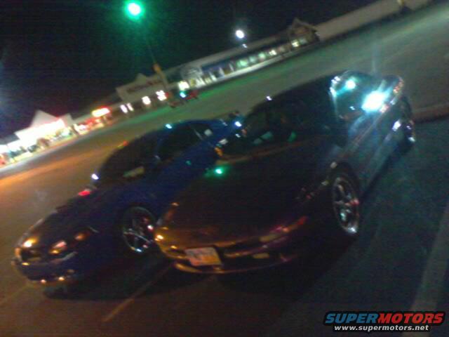 Two sports cars parked in a lot at night, one dark blue and one black. A store is visible in the background