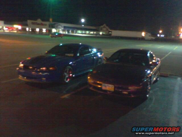Two cars parked in a lot at night. One is blue, the other is dark colored