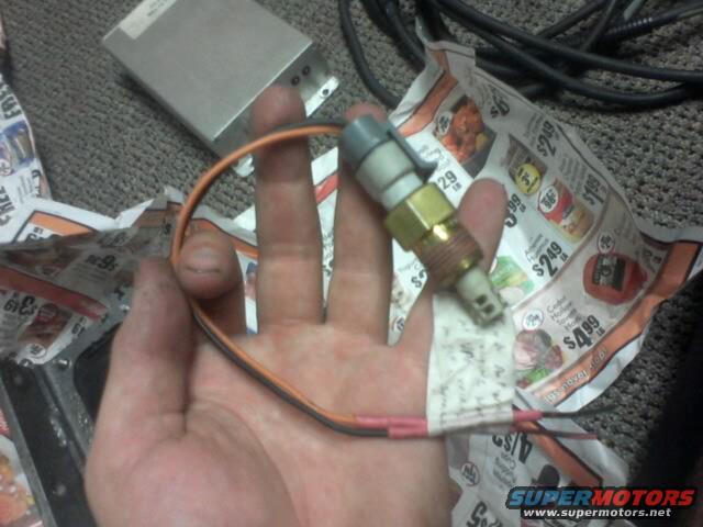 Hand holding a brass temperature sensor with orange and black wires. Newspaper ads are visible in the background