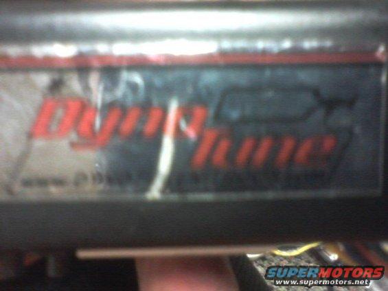 Close-up of a red "DynoTune" sticker on a dark surface, with a Supermotors logo at the bottom right