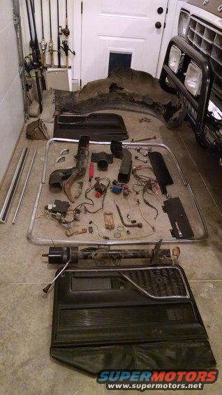 Disassembled vehicle parts including a door panel, HVAC components, and wiring, laid out on a garage floor next to fishing rods and a partial vehicle front end