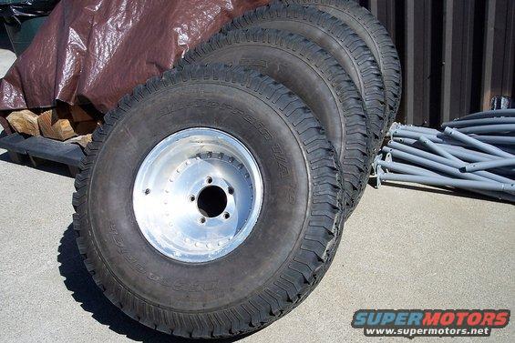 Four off-road tires with aluminum rims stacked, partially covered by a brown tarp