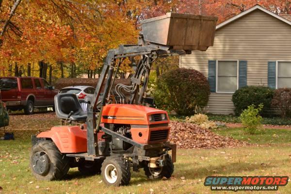 Orange tractor with front loader in yard, red SUV and white car in background