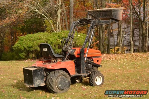 Orange tractor with a black front loader and seat, parked on a grassy lawn with fall foliage