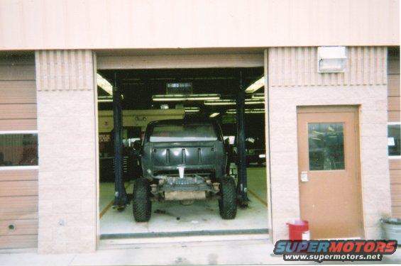 Dark-colored truck chassis on a lift inside a garage, viewed from the rear