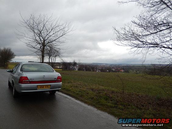 Silver sedan parked on the side of a wet road with a town in the distance under a cloudy sky