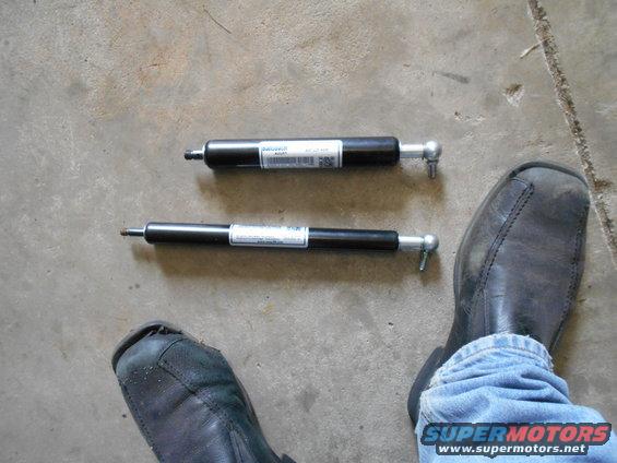 Two black gas struts with white labels and silver ball joint ends on a concrete floor, next to a person's feet in jeans and black shoes