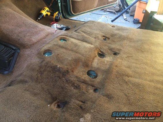 Vehicle interior floor with carpet removed, showing mounting holes and a drill