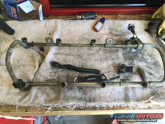 Fuel rail assembly with injectors removed, sitting on a towel with tools like a brush and utility knife