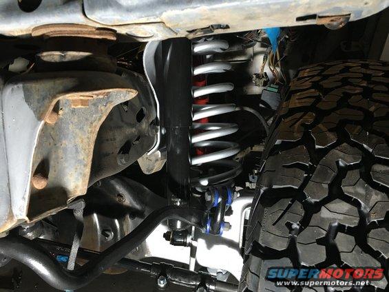 Close-up of a truck's front suspension components and a large tire. Visible text reads SUPERMOTORS.NET