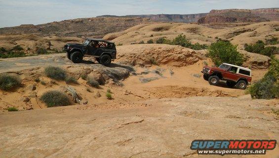Two Ford Broncos, one black and one red with white top, navigate rocky desert terrain