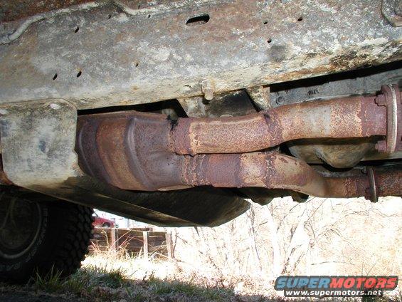 Underside view of a heavily rusted exhaust system and frame of a vehicle. A tire is visible on the left