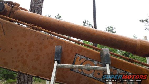 Close-up of an excavator's hydraulic cylinder and boom, showing rust and a partial logo. A ladder is visible
