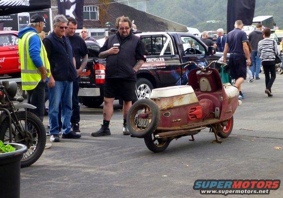 Rusty red scooter with white seat and spare tire on back, parked near a pickup truck and other people