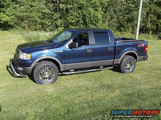 Blue Ford F-150 pickup truck parked on grass with trees in background. Has chrome wheels and side steps