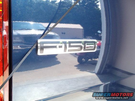Close-up of an F-150 emblem on the side of a blue truck. Another vehicle is reflected in the paint