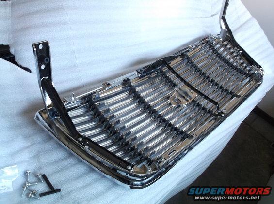 Chrome grille with horizontal bars and black accents, resting on white padding. Mounting brackets and screws are visible