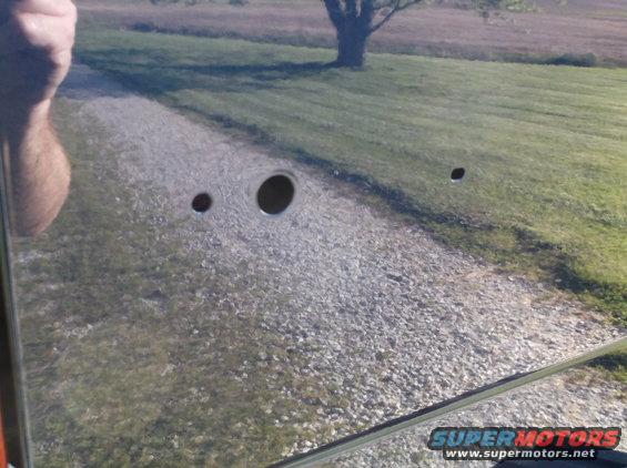 Vehicle door with three holes drilled into it. Reflection shows a gravel driveway and grassy field
