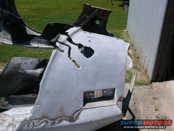 Damaged gray car bumper with large cracks and rust spots, showing a license plate mount