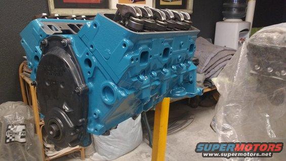 Blue V8 engine block on a stand, with valve covers visible. Another engine part is covered in plastic to the right