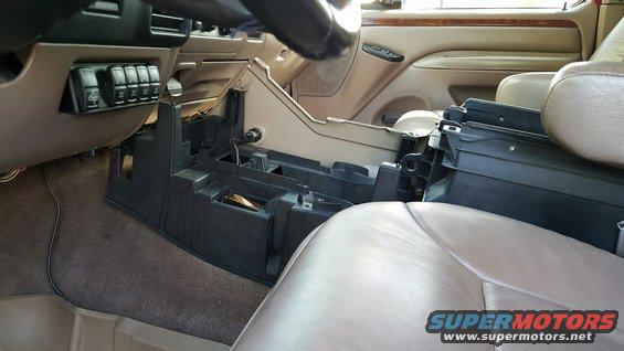 Interior of a vehicle with the center console removed, showing wiring and empty compartments. A bank of switches is visible on the left