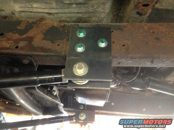 Undercarriage of a vehicle showing rusty frame and new suspension bracket with green and gold bolts