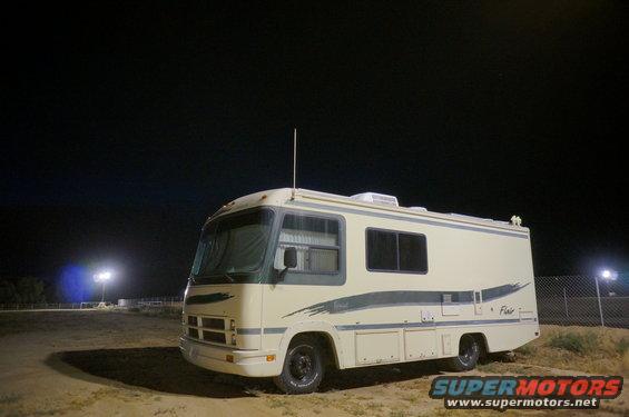 Cream-colored motorhome with dark blue stripe, parked on dirt at night. "Flair" text visible on side