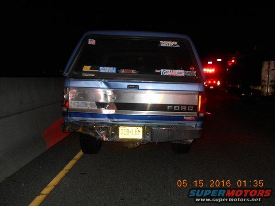 Rear of a blue Ford pickup truck with a camper shell, damaged bumper, and stickers on the back window. Another vehicle with flashing lights is visible ahead