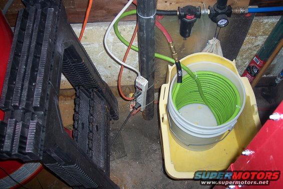 Air compressor setup with coiled green hose in a bucket, black step stool, and electrical outlet