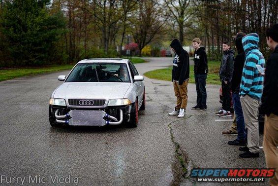 Silver 2001 Audi S4 with large front-mounted intercooler and custom piping, parked on a wet road with a group of people watching