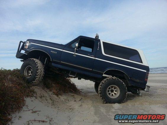 1994 Ford Bronco with large tires climbing a sandy dune near the ocean