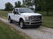 2011 Ford F-250 - A white 2011 Ford F-250 pickup truck is parked on a gravel driveway with a grassy field and trees in the background.