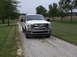 2011 Ford F-250 - A white 2011 Ford F-250 is parked on a gravel driveway surrounded by grass and trees.
