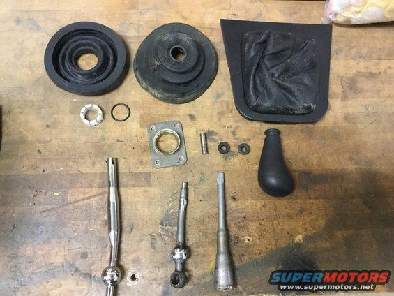 Car parts including shift knobs, boots, and levers arranged on a wooden surface. A watermark reads SUPERMOTORS.NET