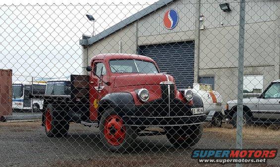 Red vintage truck with black fenders and red wheels parked in front of a building. License plate reads 91 M16