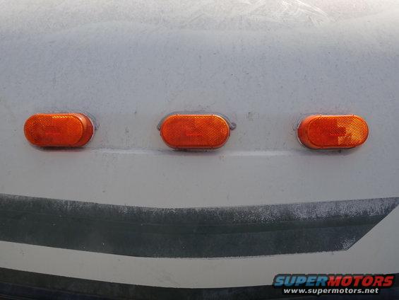Three oval orange marker lights on a white vehicle's upper body, with a dark stripe below