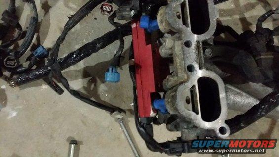 Engine intake manifold with blue-capped fuel injectors, red fuel rail, and black wiring harness laid on concrete floor