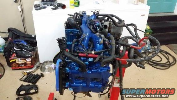 Blue Subaru engine on a red stand, with various hoses and wiring. Some tools and shoes are on the floor nearby