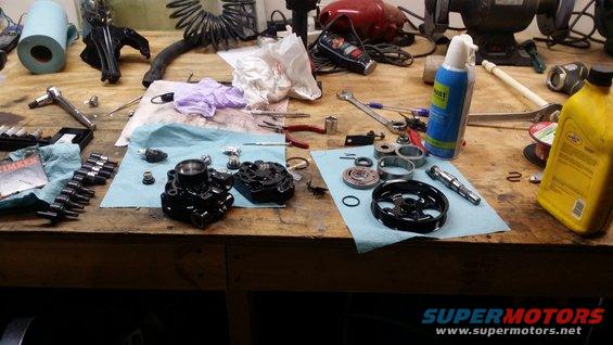 Various automotive parts, including a power steering pump and pulley, laid out on a wooden workbench with tools and cleaning supplies