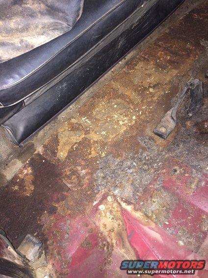 Interior floor of a vehicle with extensive rust and corrosion, showing a black seat base and red painted metal