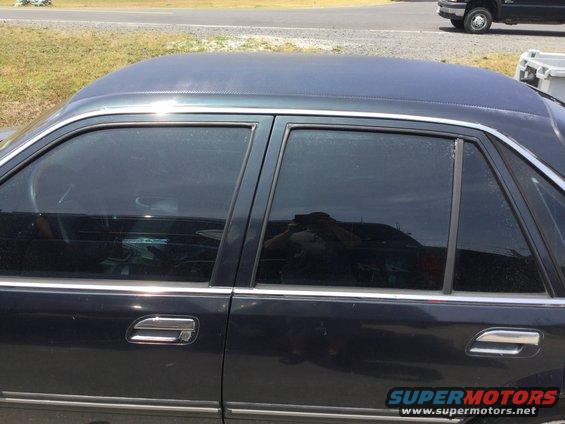 Dark sedan side view, tinted windows, chrome trim, reflection of photographer. Another vehicle visible in background