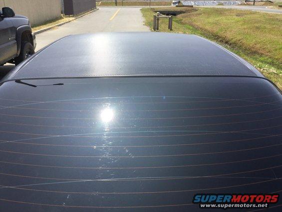 Dark colored car roof and rear window with defroster lines. A black pickup truck is parked to the left