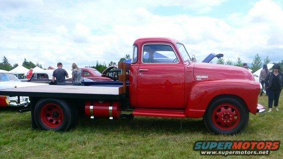 Red flatbed truck with wooden sides, seen from the side. Two people sit on the bed