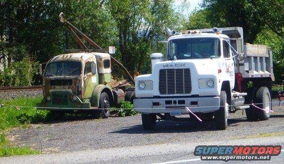 Front view of a rusty old truck with a crane next to a white Mack dump truck