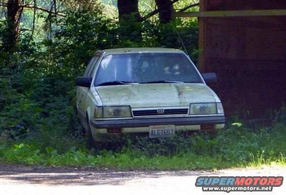 Front view of a dusty, light-colored Subaru sedan parked in overgrown grass next to a wooden structure. License plate reads ALG1607